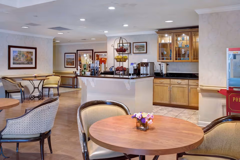 A cozy dining area in a senior living facility featuring round wooden tables with cushioned chairs. There is a counter with a coffee and beverage station, including dispensers and a tiered fruit basket. The background shows wooden cabinets with glass doors displaying dishes, framed artwork on the walls, and a popcorn machine on the right side.