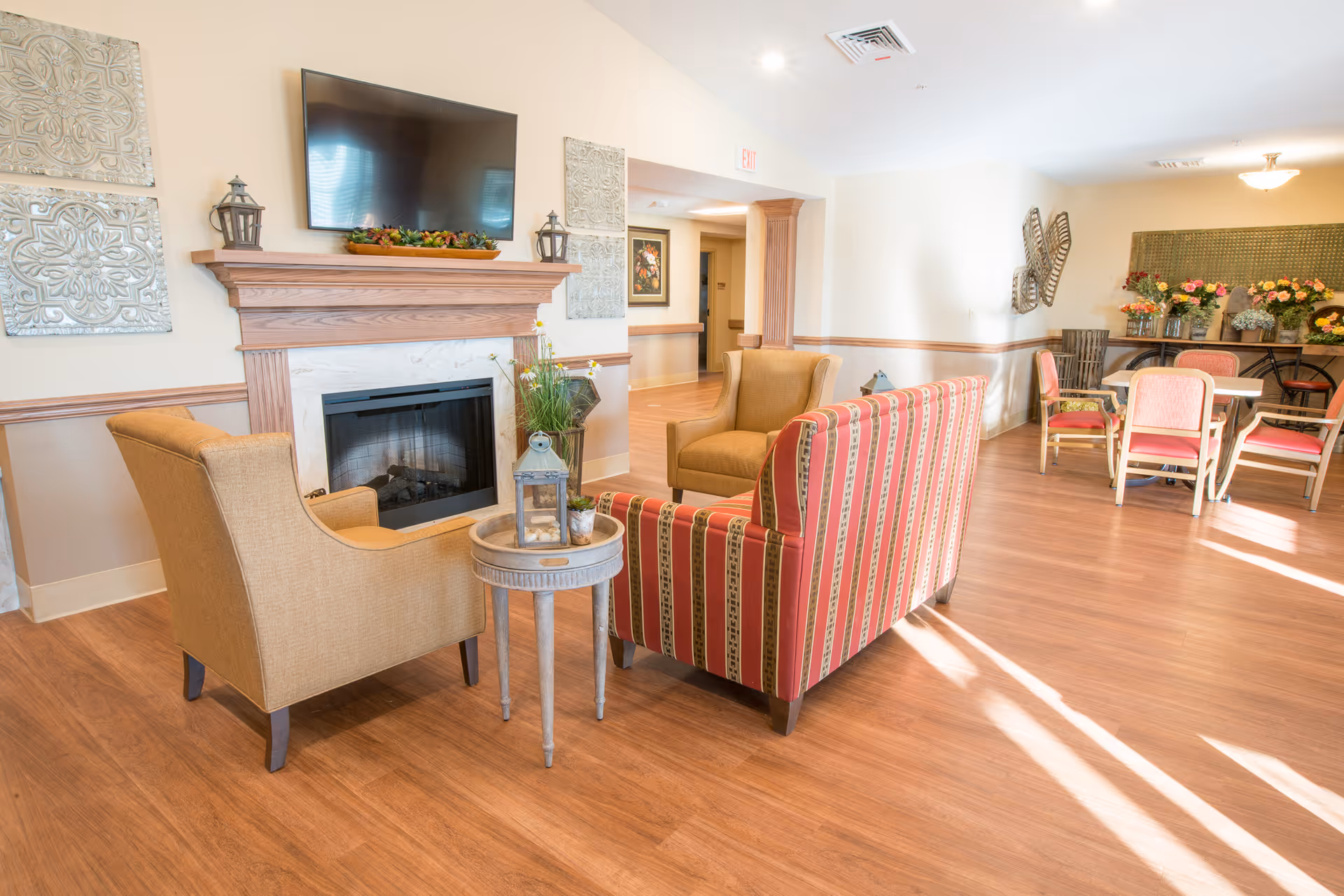 Bright lounge with armchairs and a striped sofa arranged around a fireplace and TV, with dining tables visible in the background.