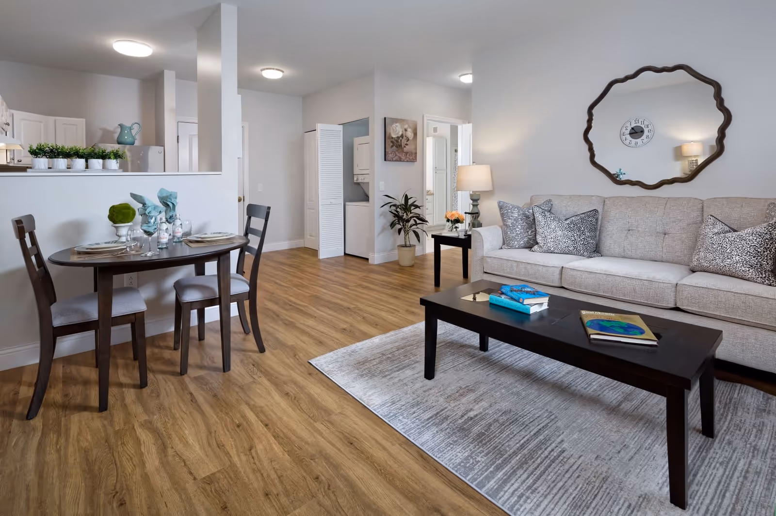 Bright living room with a beige sofa, coffee table, small dining table, and an open kitchen in the background.