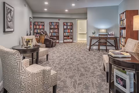 A cozy interior common area with patterned carpet, two upholstered armchairs with a small round table between them, bookshelves filled with books along the back wall, a brown recliner chair, and a wooden desk with a chair and a lamp. The walls are painted light gray and blue, and there is framed artwork on the wall.