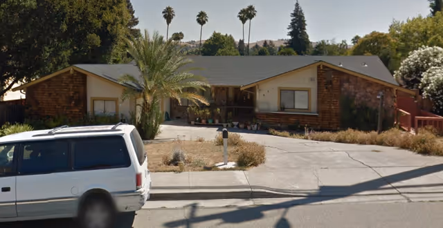Single-story residential building with a sloped roof and a mix of wood and stone exterior. There is a driveway and a white van parked in front. Palm trees and other greenery surround the property under a clear sky.