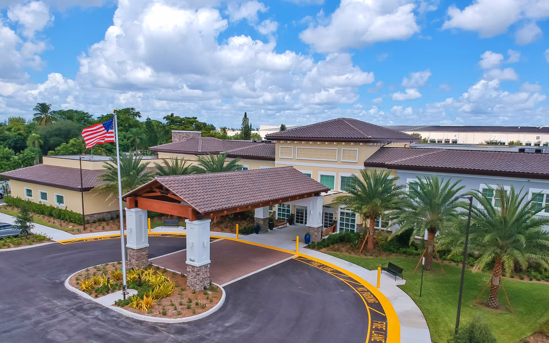 Exterior view of a senior living facility with a covered entrance, palm trees, an American flag, and a circular driveway under a partly cloudy sky.