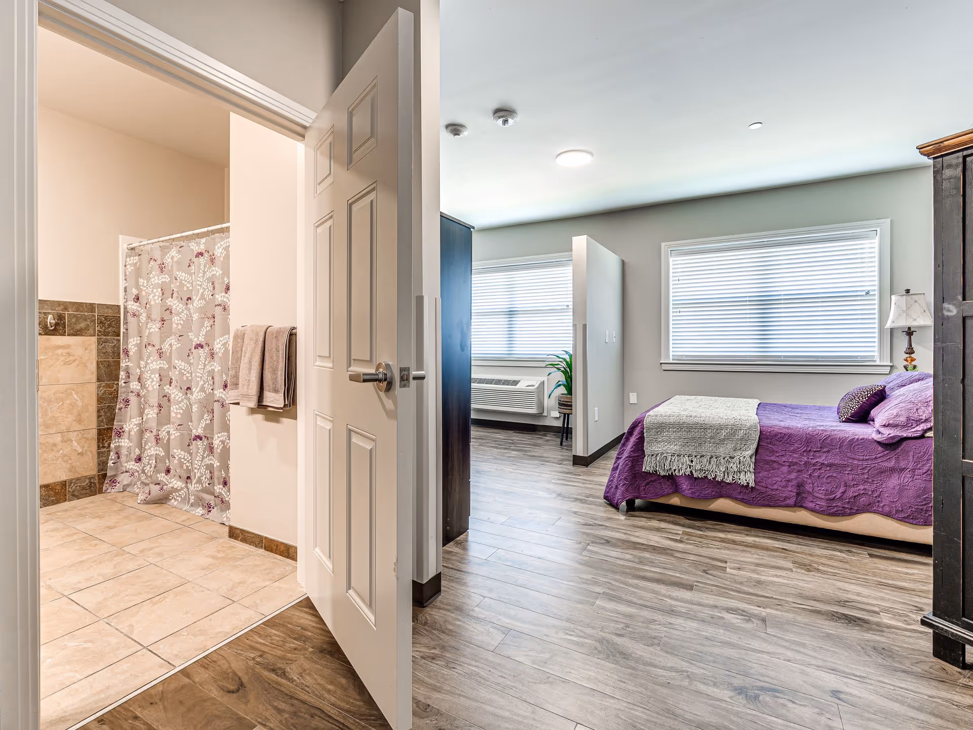 View of a bedroom with a purple bedspread and pillows, a window with blinds, a dark wooden cabinet, and a small lamp. The bedroom has wood flooring and is adjacent to a bathroom with beige tiled floor and walls, a shower with a floral curtain, and two towels hanging on a rack.