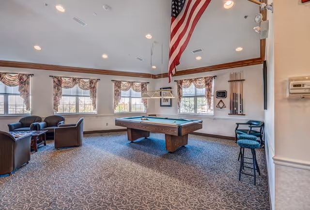 A spacious recreational room with a pool table in the center, surrounded by several windows with patterned valances. There are comfortable seating areas with armchairs and a small table on the left side. Pool cues and a rack are mounted on the wall to the right, along with two bar stools. An American flag hangs from the ceiling above the pool table. The room has carpeted flooring and recessed lighting.