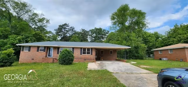 Single-story brick house with a carport and a driveway, surrounded by green grass and trees under a partly cloudy sky. A blue vehicle is partially visible on the right side of the image.
