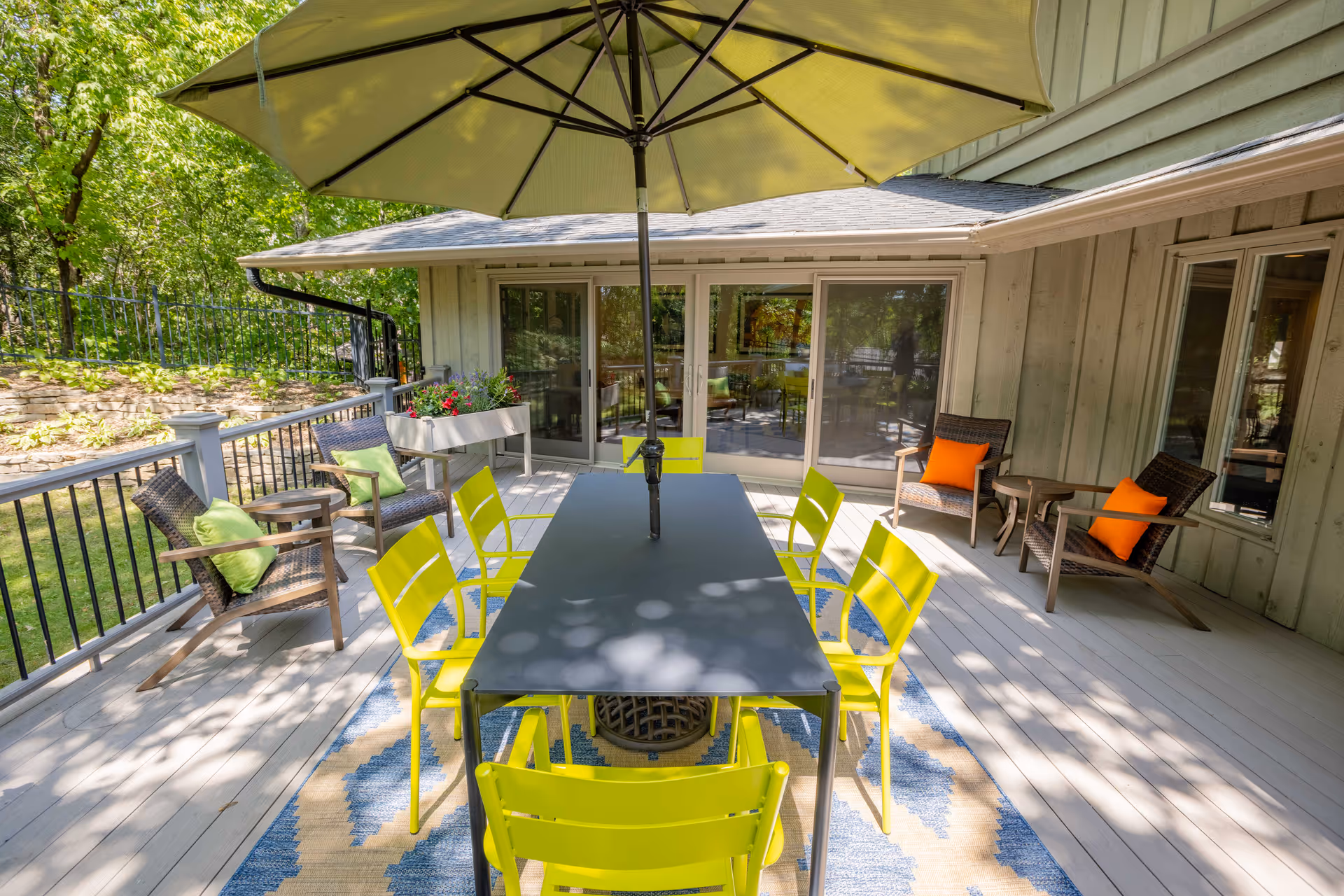 Sunlit outdoor deck with a rectangular dining table under a large umbrella, bright yellow chairs, and lounge seating by sliding glass doors.