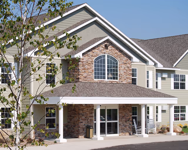 Exterior front view of a senior living facility building with beige and stone facade, large windows, a covered entrance supported by white columns, and a small tree in the foreground.