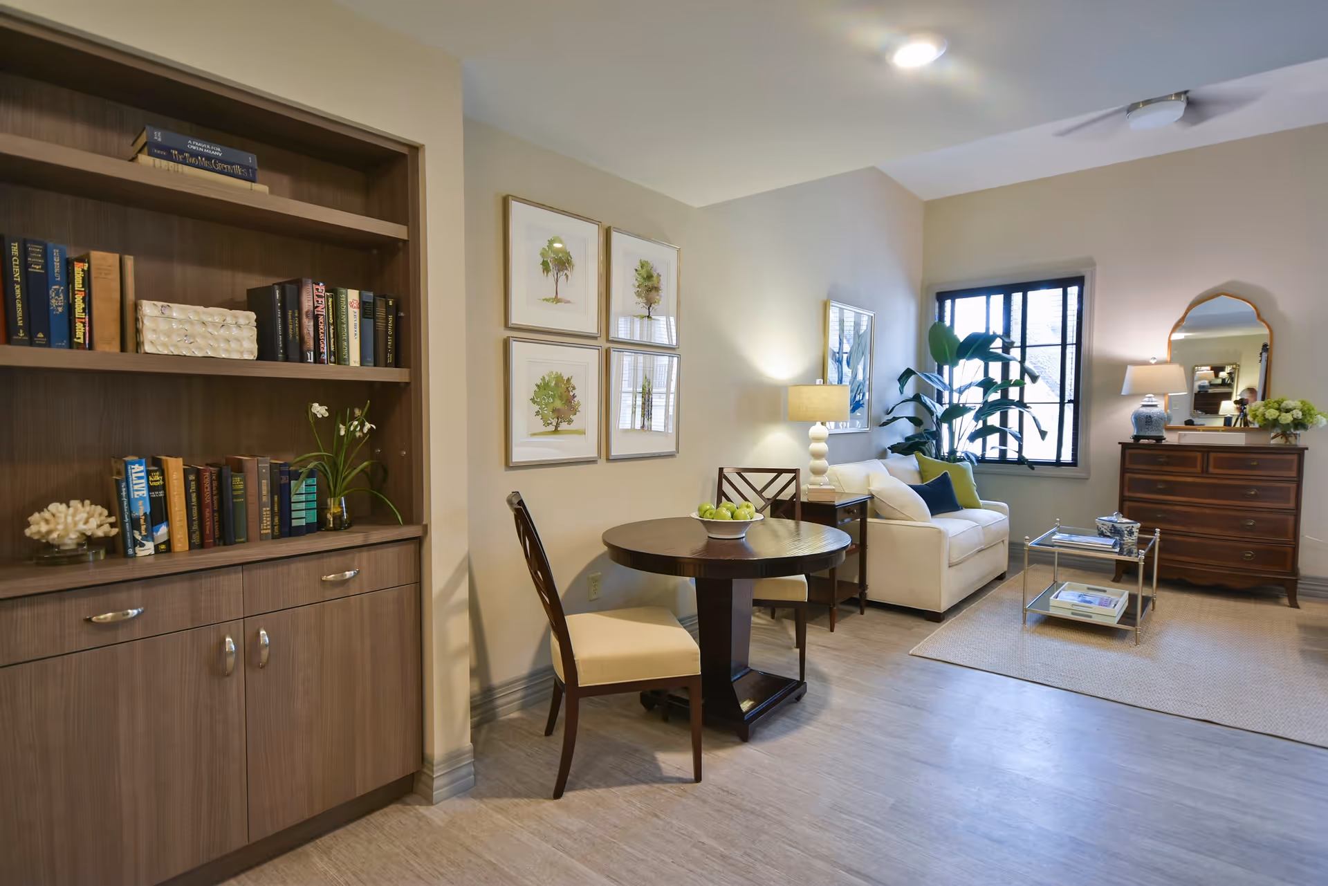 Sunlit living room with a round dining table and chairs, a sofa by a window, bookshelves and a dresser with decorative accents.