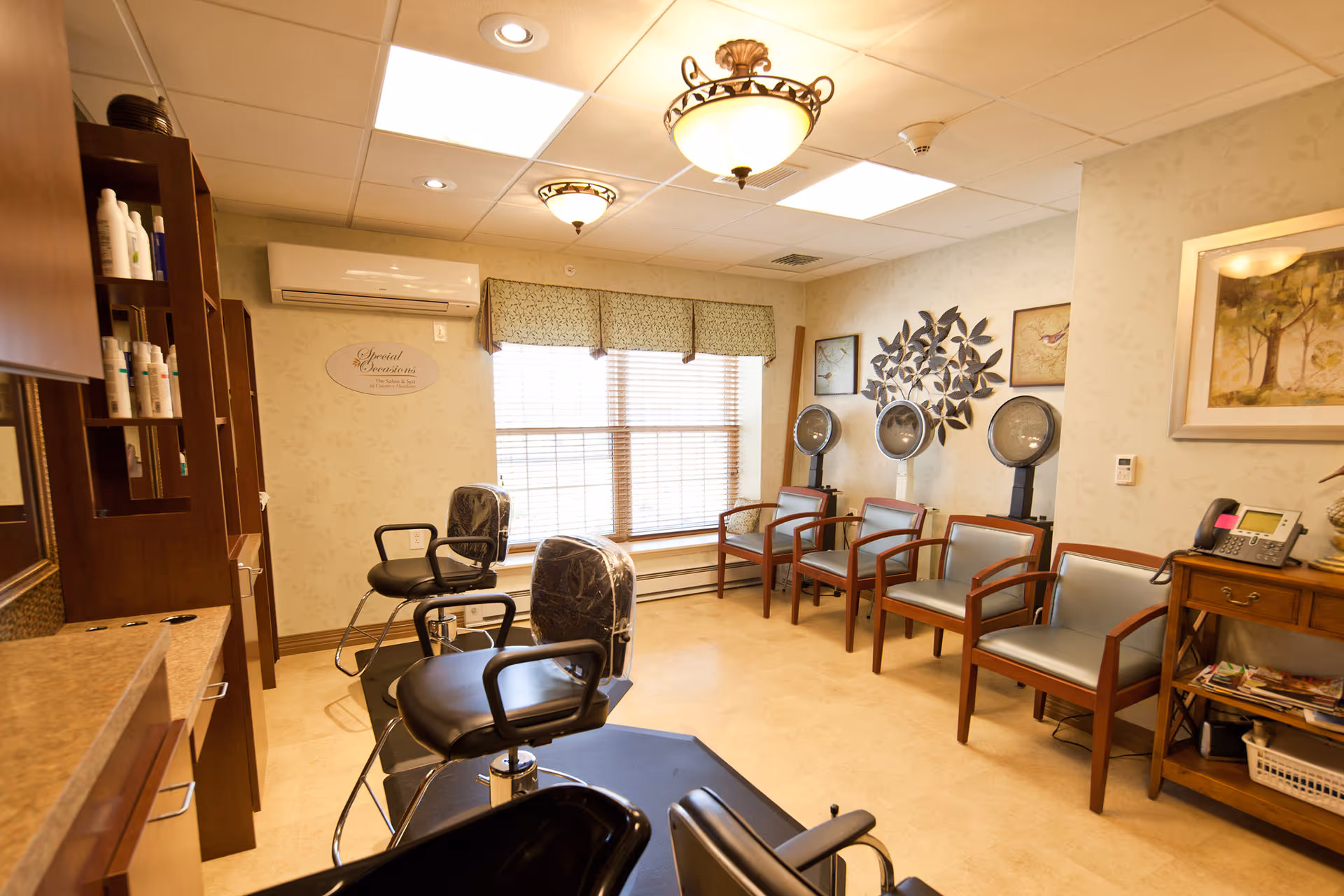 Interior of a hair salon area in a senior living facility with salon chairs, hair dryers, wooden shelving with hair products, and seating along the wall. The room has a large window with blinds and valance, decorative wall art, and warm lighting.