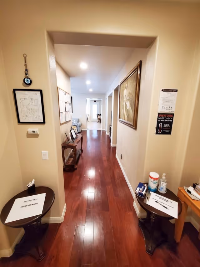 A hallway with polished wooden floors and beige walls, featuring framed artwork and notices on the walls. Two small tables are positioned on either side of the hallway entrance, each holding visitor sign-in sheets and hand sanitizer. The hallway leads to a well-lit room with a window at the far end.