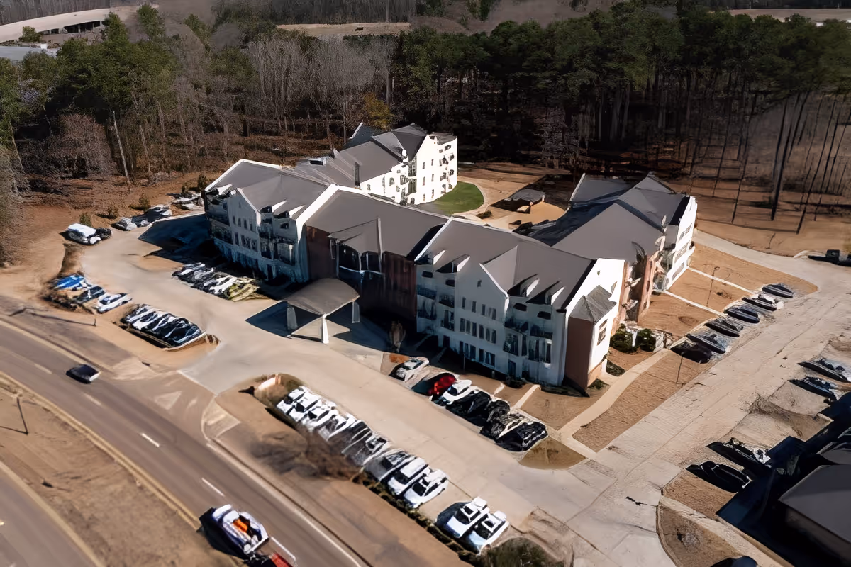 Aerial view of SummerHouse Beau Ridge, a multi-story senior living facility surrounded by parking lots and wooded areas. The building has a light-colored exterior with multiple peaked roofs and a covered entrance. Several cars are parked in the parking areas around the building.