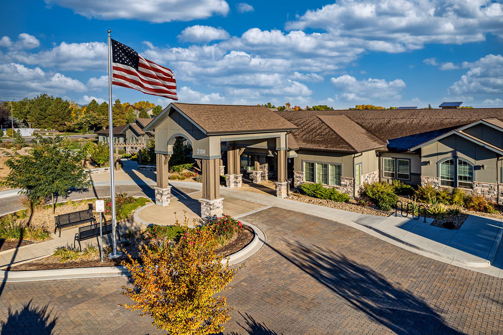Exterior view of Aspens at Fort Collins Memory Care facility showing the entrance with a covered driveway, an American flag on a flagpole, landscaped gardens, benches, and a clear blue sky with scattered clouds.