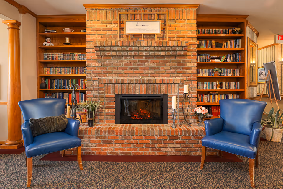 A cozy interior living area featuring a large brick fireplace flanked by built-in wooden bookshelves filled with books and decorative items. Two blue leather armchairs with wooden legs are positioned on either side of the fireplace, with a small plant and candle holders placed on the hearth. The room has warm lighting and a patterned carpet floor.