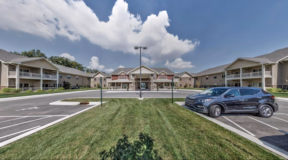 Front exterior of a two-story senior living building with a covered main entrance, parking lot, and landscaped lawn under a partly cloudy sky.