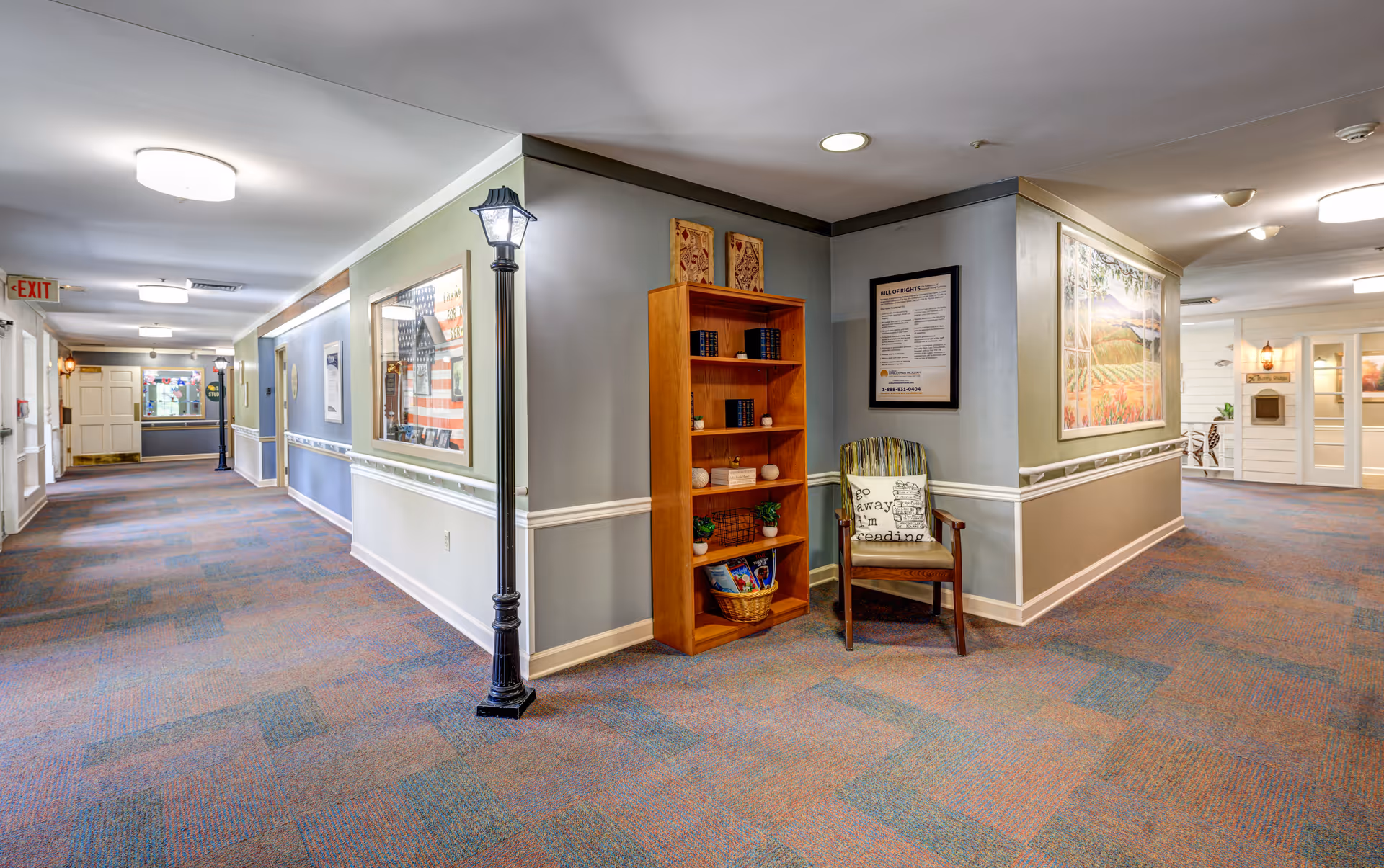 A well-lit hallway in a senior living facility with carpeted floors and light-colored walls. There is a wooden bookshelf filled with books and decorative items, next to a wooden chair with a cushion that reads 'go away I'm reading.' The hallway features framed artwork and informational posters on the walls, with a lamp post-style light fixture near the bookshelf. Doors and exit signs are visible further down the corridor.