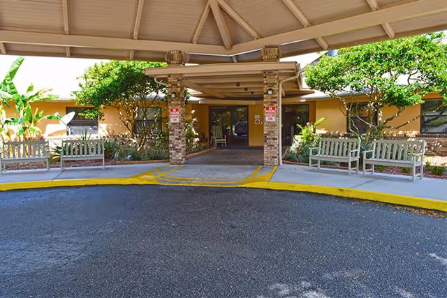 Covered entrance to a building with a circular driveway, benches on either side, and greenery including trees and bushes around the entrance.