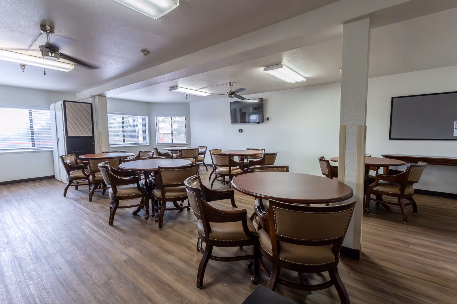 A bright and spacious common dining area with multiple round wooden tables and cushioned chairs arranged neatly. The room has large windows letting in natural light, a wall-mounted TV, ceiling fans, and wood-style flooring.