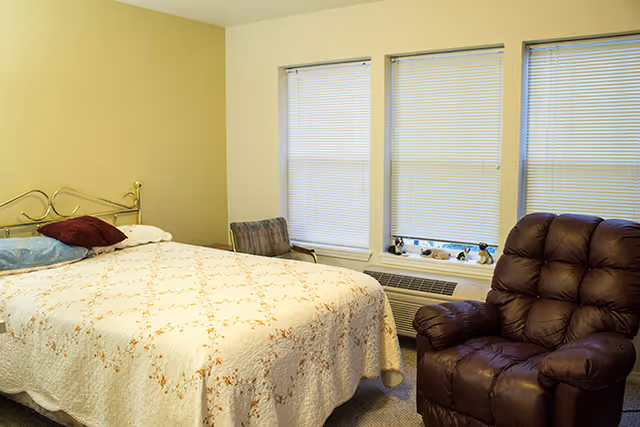 A senior living bedroom with a bed covered in a white floral quilt, a brown leather recliner chair, a small striped armchair, and three windows with closed blinds above a heating unit.