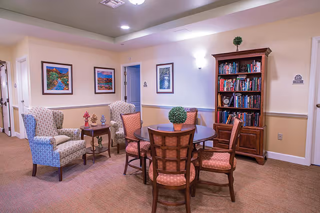 A cozy common area in a senior living facility featuring a round wooden table with four cushioned chairs around it. There is a potted plant centerpiece on the table. To the left, there are two upholstered armchairs with a small wooden side table between them, decorated with small figurines. On the right side, there is a tall wooden bookshelf filled with books and decorative items. The walls are painted in light beige with white trim, and three framed landscape paintings hang on the walls. The carpet is a neutral brown color, and the ceiling has recessed lighting.