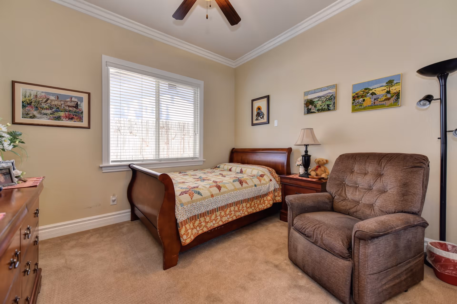 A cozy bedroom with a wooden sleigh bed covered with a patchwork quilt, a brown upholstered armchair, a wooden nightstand with a lamp and a teddy bear, a wooden dresser with framed photos and flowers, and several framed paintings on beige walls. A window with white blinds lets in natural light.