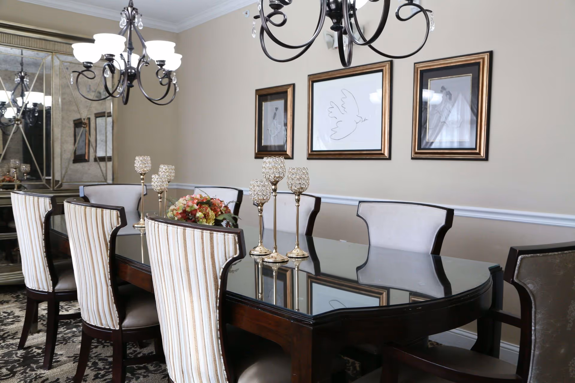 A formal dining room with a large dark wood table topped with a reflective glass surface. The table is surrounded by eight chairs, some with striped upholstery and others with plain light fabric. On the table are decorative candle holders and a floral centerpiece. The walls are beige with white trim, decorated with three framed pictures, including one of a dove. A chandelier with multiple lights hangs above the table, and a large mirror is visible on one side of the room.