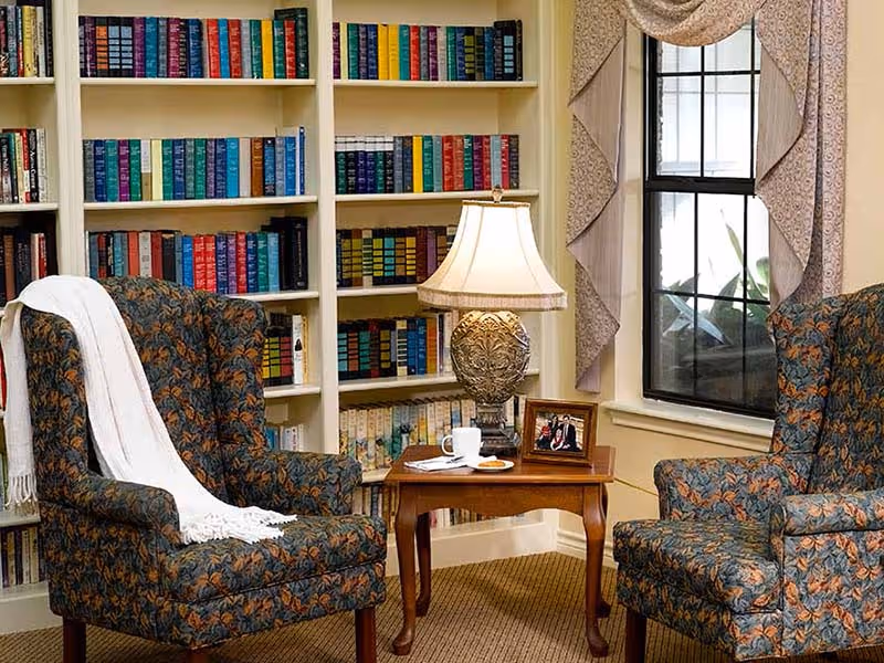 Cozy sitting area with two patterned armchairs, a small wooden side table holding a lamp and framed photo, and bookshelves behind a window.