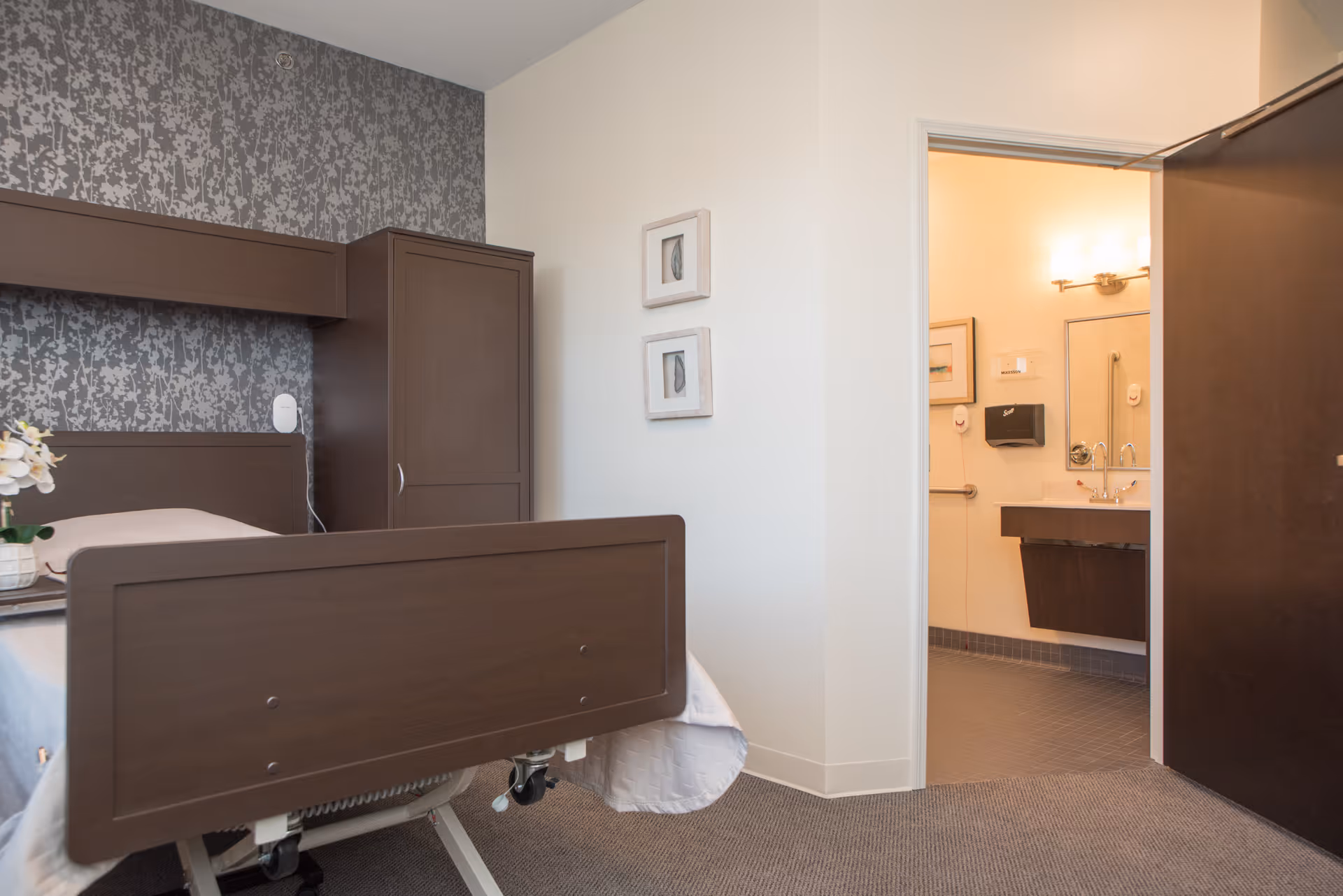 A senior living facility bedroom with a hospital-style bed, dark wood furniture including a wardrobe and headboard, floral wallpaper behind the bed, and an open door leading to a bathroom with a sink, mirror, and handrails.