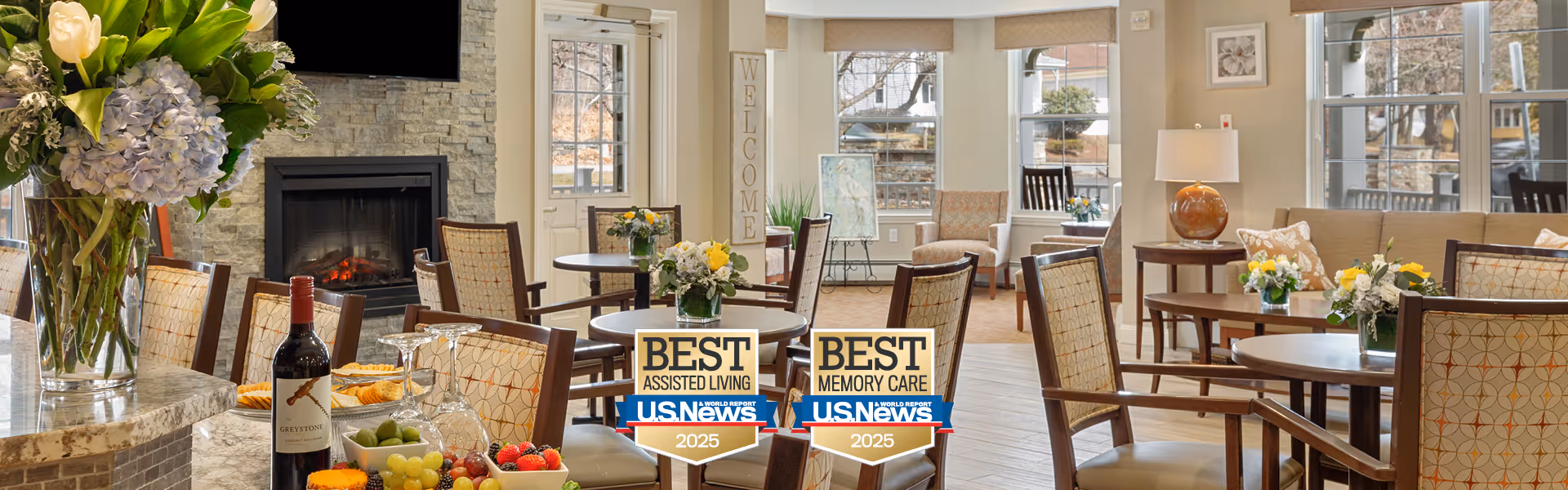 A bright and welcoming dining area in a senior living facility with multiple round tables and chairs, floral centerpieces on the tables, a fireplace with a TV mounted above it, large windows letting in natural light, and a bottle of wine with glasses on a granite countertop.
