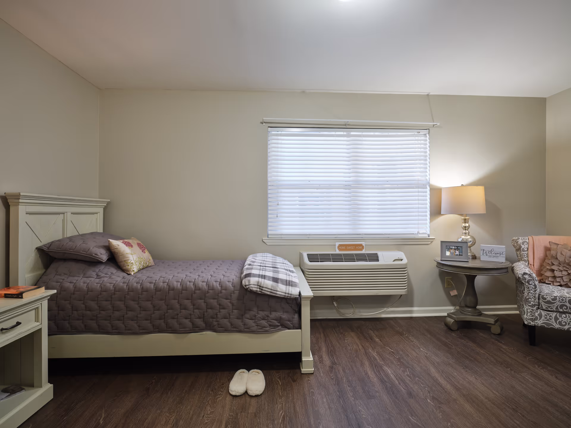 A cozy bedroom with a single bed covered in a gray quilt and decorative pillows. Next to the bed is a small nightstand with a book on it. A window with closed blinds is above a wall-mounted air conditioning unit. To the right, there is a round side table with a lamp, a framed photo, and a welcome sign. An upholstered armchair with a floral pattern and a pink throw pillow is partially visible. A pair of white slippers is placed on the wooden floor near the bed.