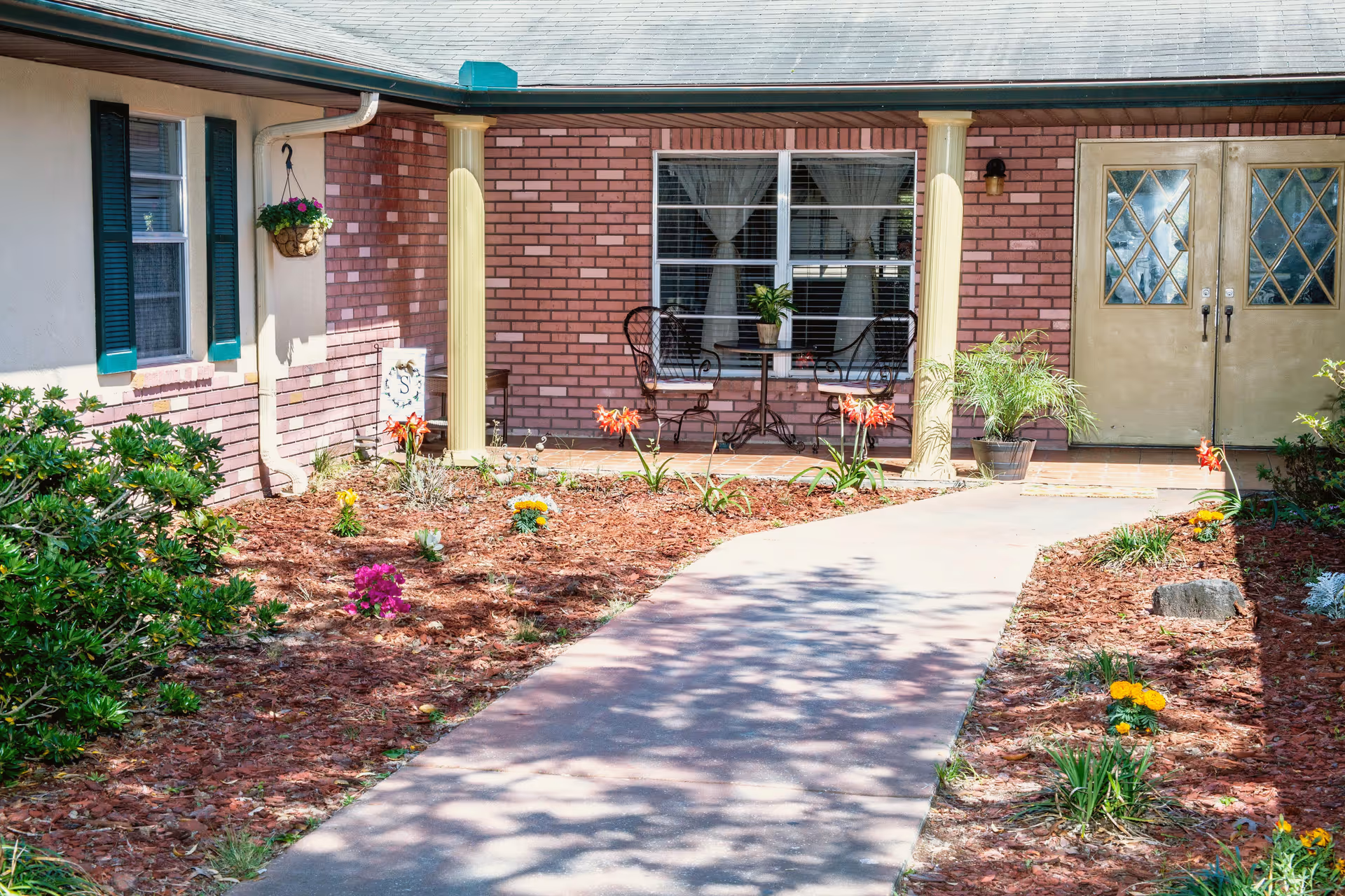 Front entrance of a building with a brick facade and beige double doors with diamond-patterned glass. There is a small patio area with two black metal chairs and a round table with a potted plant. The walkway leading to the entrance is bordered by a garden with various flowers and plants. The building has green shutters on the windows and a hanging flower basket on the left side.