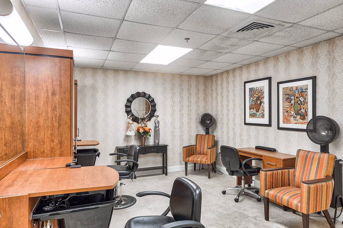 Interior view of a salon area in a senior living facility with wooden counters, black salon chairs, two hair drying stations with striped orange armchairs, a small black table with decorative items and a round mirror, and two framed floral paintings on the wall.