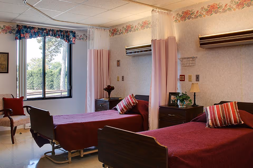 A shared bedroom in a senior living facility with two hospital-style beds covered in maroon bedspreads and striped pillows. Each bed has a wooden nightstand with a lamp, a plant, and a framed photo. Pink privacy curtains hang between the beds. A window with floral valance lets in natural light, and a cushioned chair with a red pillow is positioned near the window. The walls have a floral border near the ceiling.