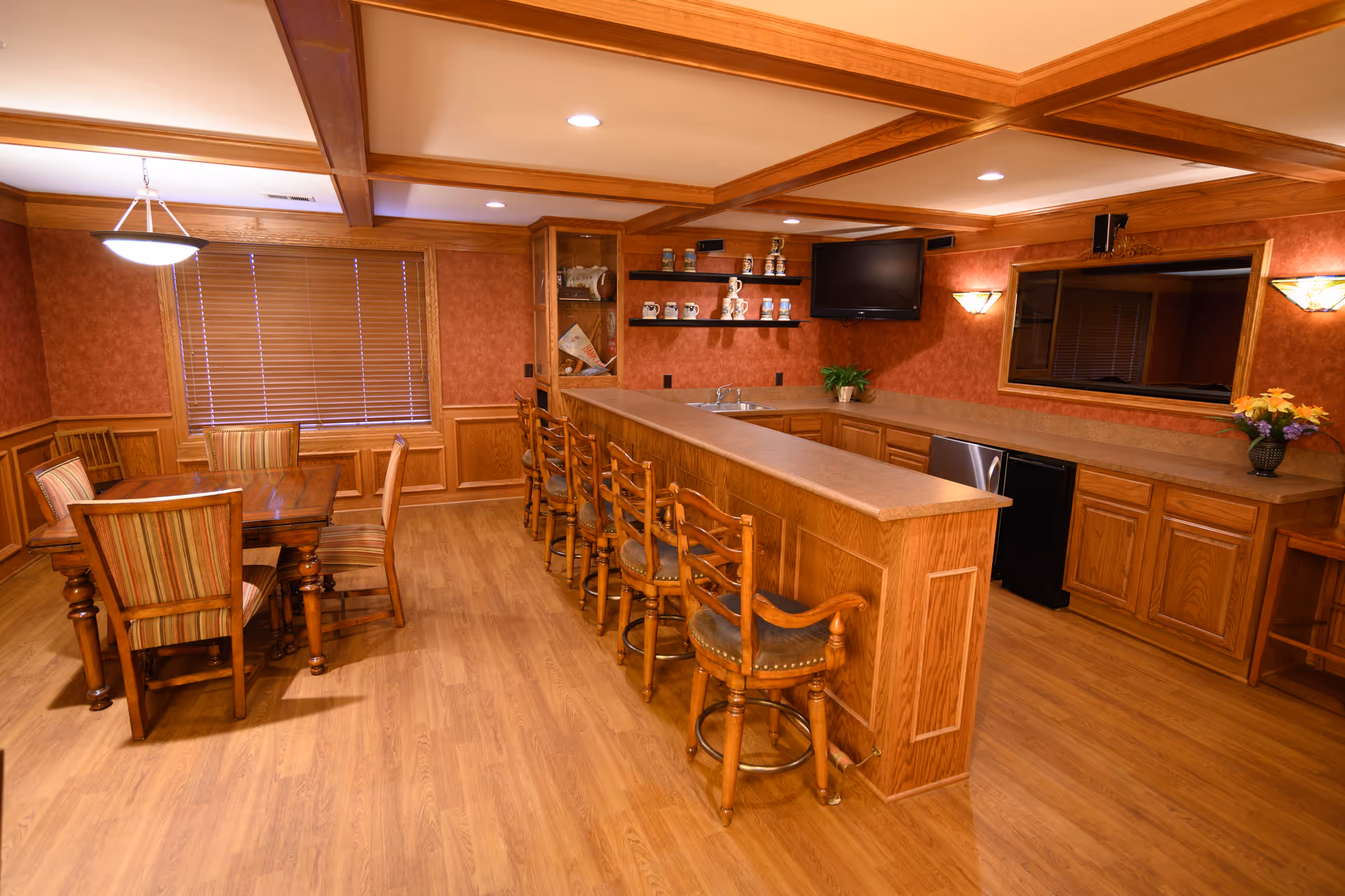 A warm, wood-paneled room featuring a long bar counter with six wooden bar stools, a dining table with six striped upholstered chairs, a flat-screen TV mounted on the wall, shelves with decorative items, and a large mirror. The room has wooden flooring, recessed lighting, and a window with closed blinds.
