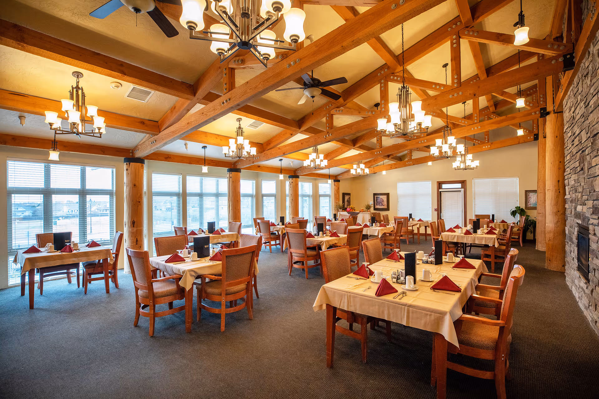 Spacious dining room with exposed wooden beams, multiple tables set with napkins and place settings, and large windows along one wall.