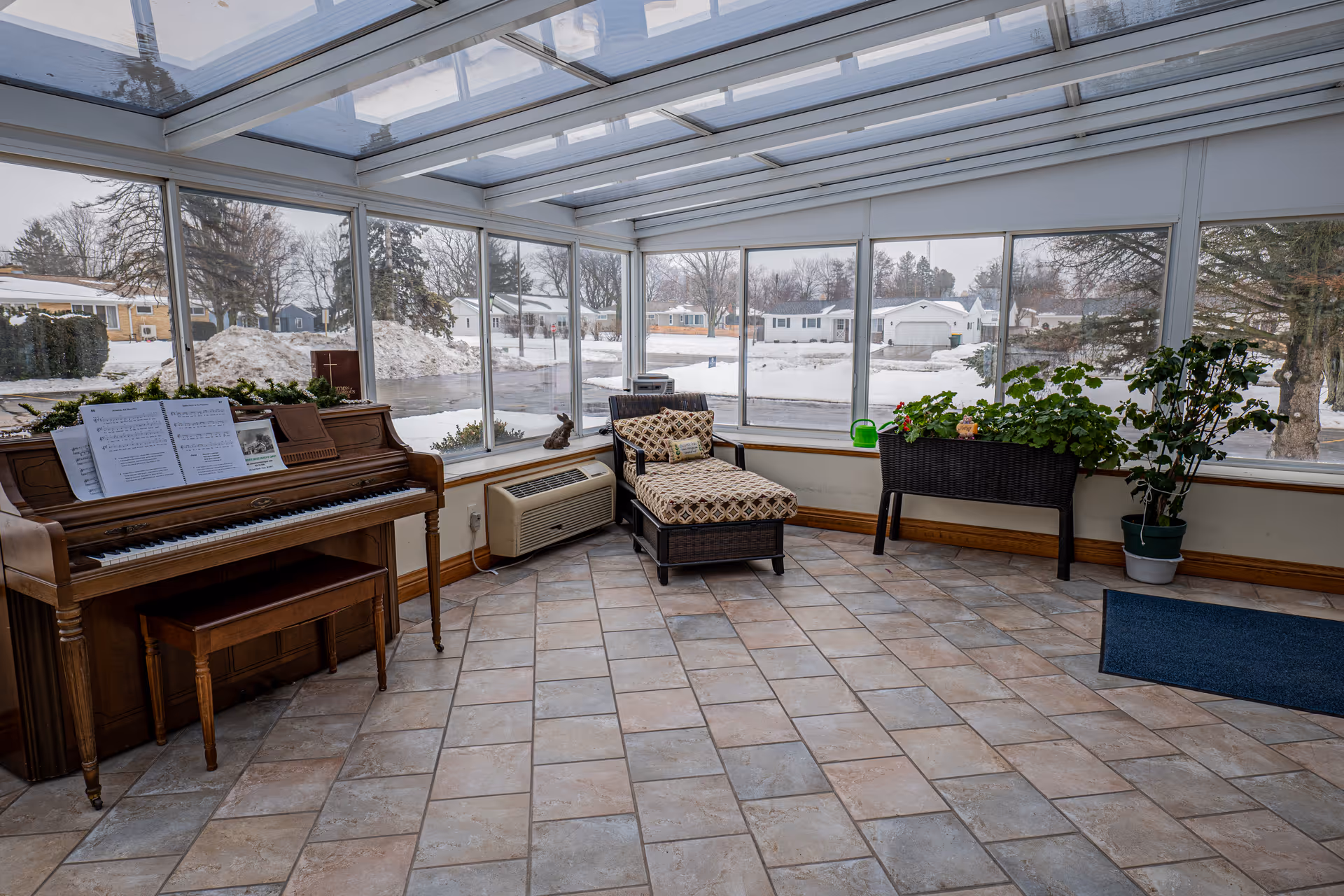 A sunroom with large windows and a glass ceiling showing a snowy outdoor scene. Inside, there is a wooden piano with sheet music on it, a cushioned wicker chaise lounge, two planters with green plants, and a blue floor mat near the entrance.