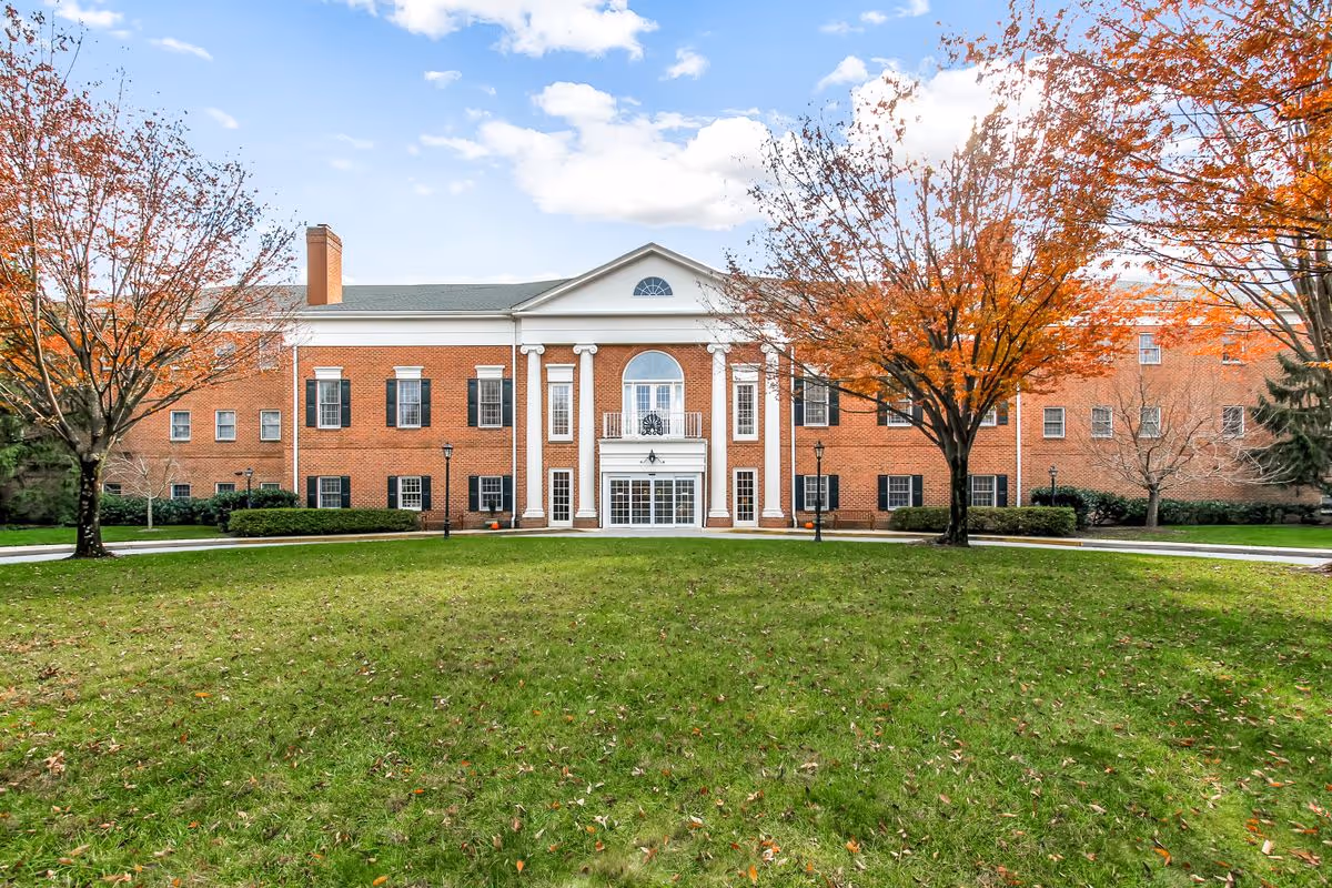 Brick building with a white-columned central entrance, flanked by autumn trees and a wide green lawn.