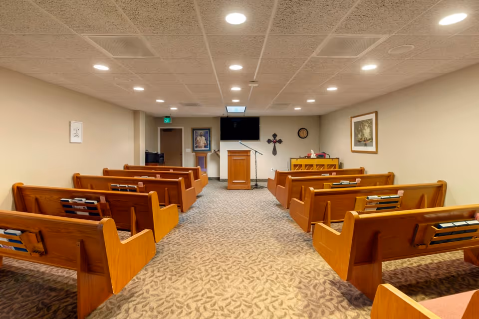 Interior view of a small chapel or meeting room with wooden pews arranged in rows facing a wooden podium with a microphone. The room has beige walls, a carpeted floor with a leaf pattern, a wall-mounted TV above the podium, a cross on the wall, a clock, and framed pictures.