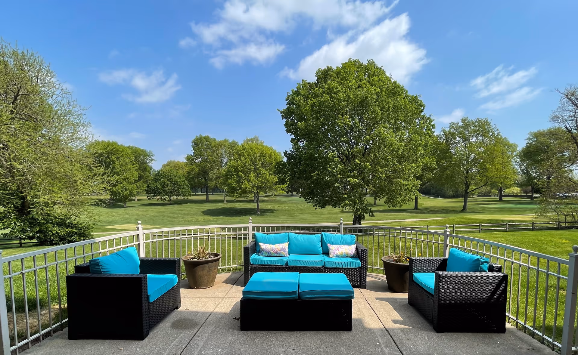 Outdoor patio area with black wicker furniture featuring bright turquoise cushions, including a sofa, two armchairs, and two ottomans. The patio is enclosed by a white metal railing and overlooks a large green lawn with several trees under a blue sky with scattered clouds.
