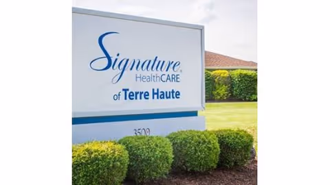 A freestanding sign that reads "Signature HealthCARE of Terre Haute" set in front of trimmed shrubs and a lawn with the building roof visible behind.