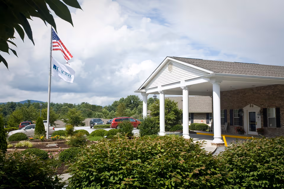 Exterior view of The Laurels of Hendersonville facility entrance with a white columned portico, landscaped bushes, and two flags on a flagpole including an American flag and a facility flag, with parked cars and trees in the background under a partly cloudy sky.