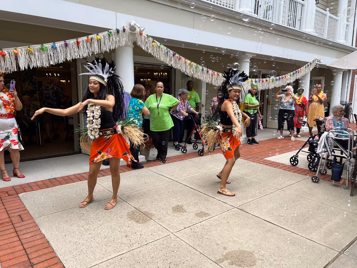 Two dancers in tropical costumes perform in a decorated senior living courtyard while residents and staff watch.
