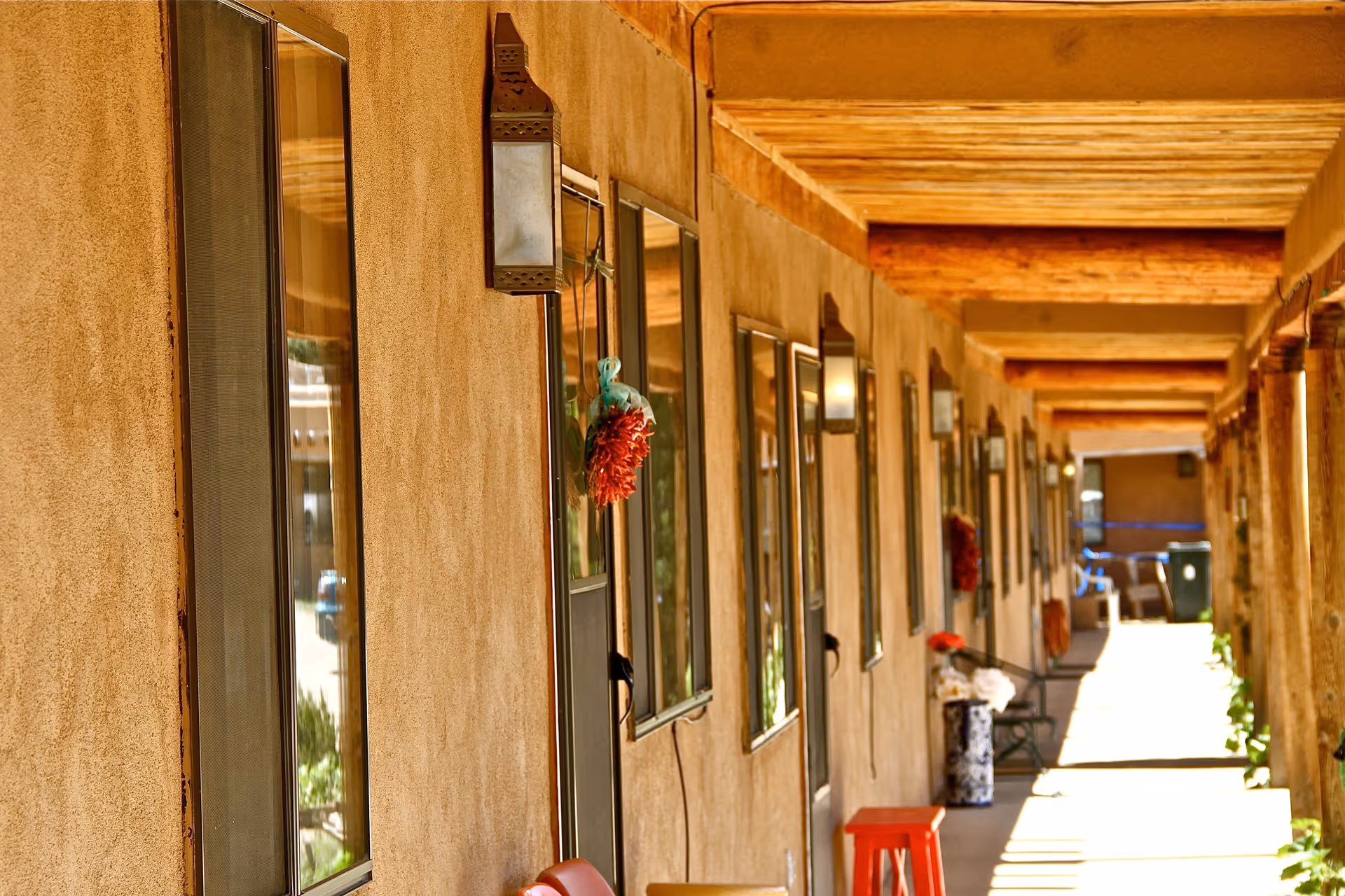 A long covered outdoor corridor with adobe-style walls and wooden beams overhead. Several windows and doors line the wall, each decorated with hanging red chili ristras. There are benches, a red stool, and potted plants along the walkway, with sunlight casting shadows on the ground.