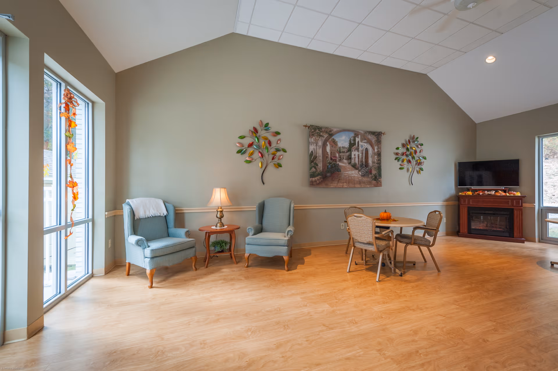 A spacious and well-lit living room area with two blue armchairs and a small wooden side table with a lamp between them. There is a round table with four chairs, a flat-screen TV on a wooden stand with a fireplace below it, and large windows letting in natural light. The walls are decorated with a painting and two metal leaf wall art pieces.