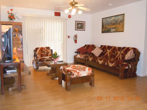 A living room with wooden furniture including a sofa, armchair, ottoman, coffee table, and TV stand. The sofa and armchair have brown and orange patterned upholstery. There is a ceiling fan with lights, a fire extinguisher mounted on the wall, a framed painting, and vertical blinds covering a sliding glass door with an exit sign above it. The floor is light wood.