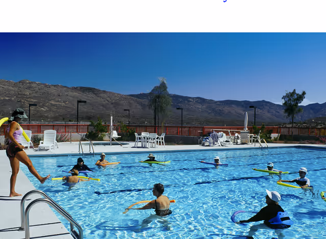 Outdoor swimming pool with several adults participating in a water exercise class using pool noodles. The pool area is surrounded by lounge chairs, tables, and a fence with mountains visible in the background under a clear blue sky.