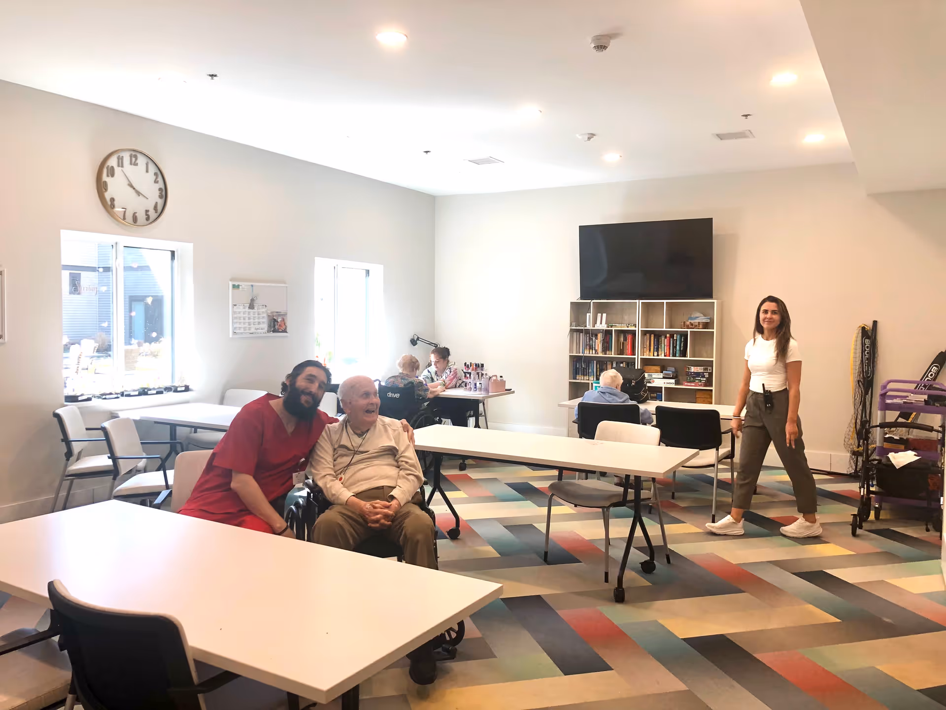 A bright common room in a senior living facility with several elderly residents and staff. One staff member in red scrubs is sitting next to an elderly man in a wheelchair, both smiling. Another staff member in casual attire is standing nearby. In the background, two elderly women are seated at a table engaged in an activity. The room has a colorful patterned floor, white walls, a large wall-mounted TV, and a bookshelf filled with books and games.