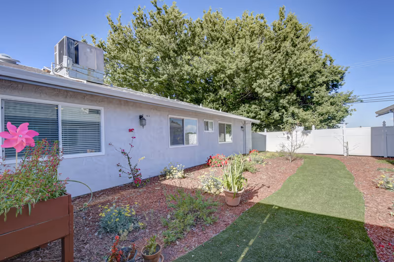 Outdoor garden area at Cherry Blossom Assisted Living with a pathway of green grass bordered by mulch and various plants and flowers. The side of a single-story building with windows and an air conditioning unit on the roof is visible. Tall trees and a white fence enclose the area under a clear blue sky.