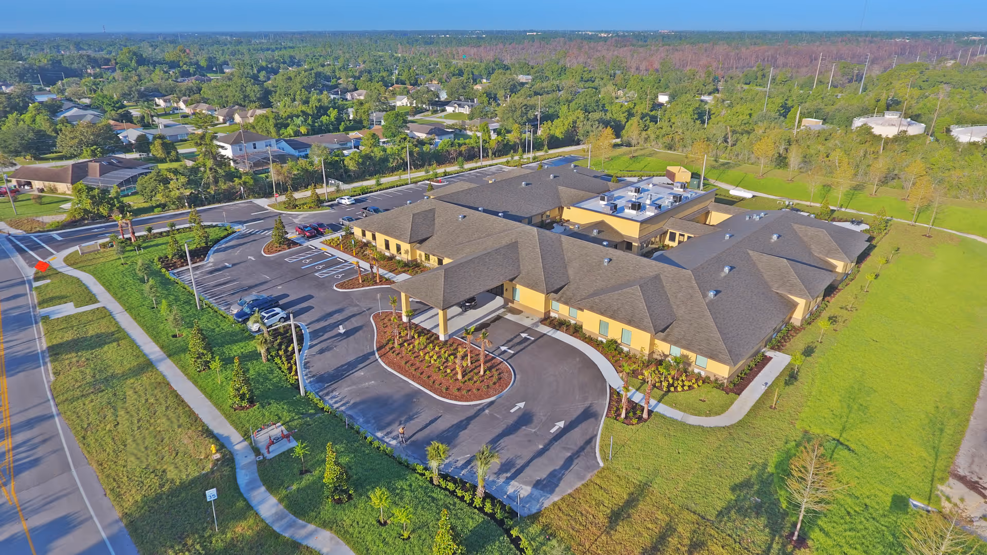 Aerial view of a single-story senior living facility named Gold Choice ALF surrounded by greenery and residential houses. The building has a large parking lot with several cars parked, landscaped areas with palm trees and shrubs, and a covered entrance driveway. The surrounding area includes sidewalks, roads, and open grassy spaces.