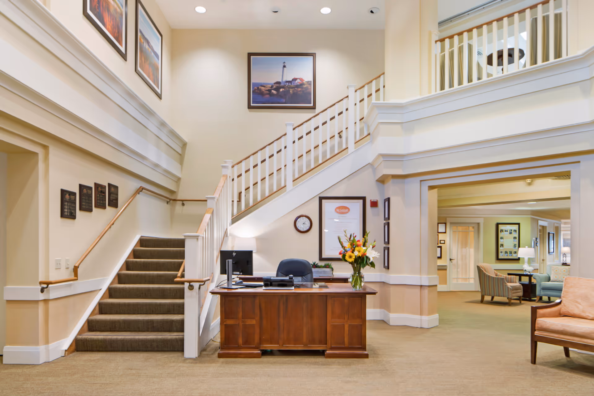 Reception area inside a senior living facility featuring a wooden front desk, a staircase with white railings, and a seating area to the right.