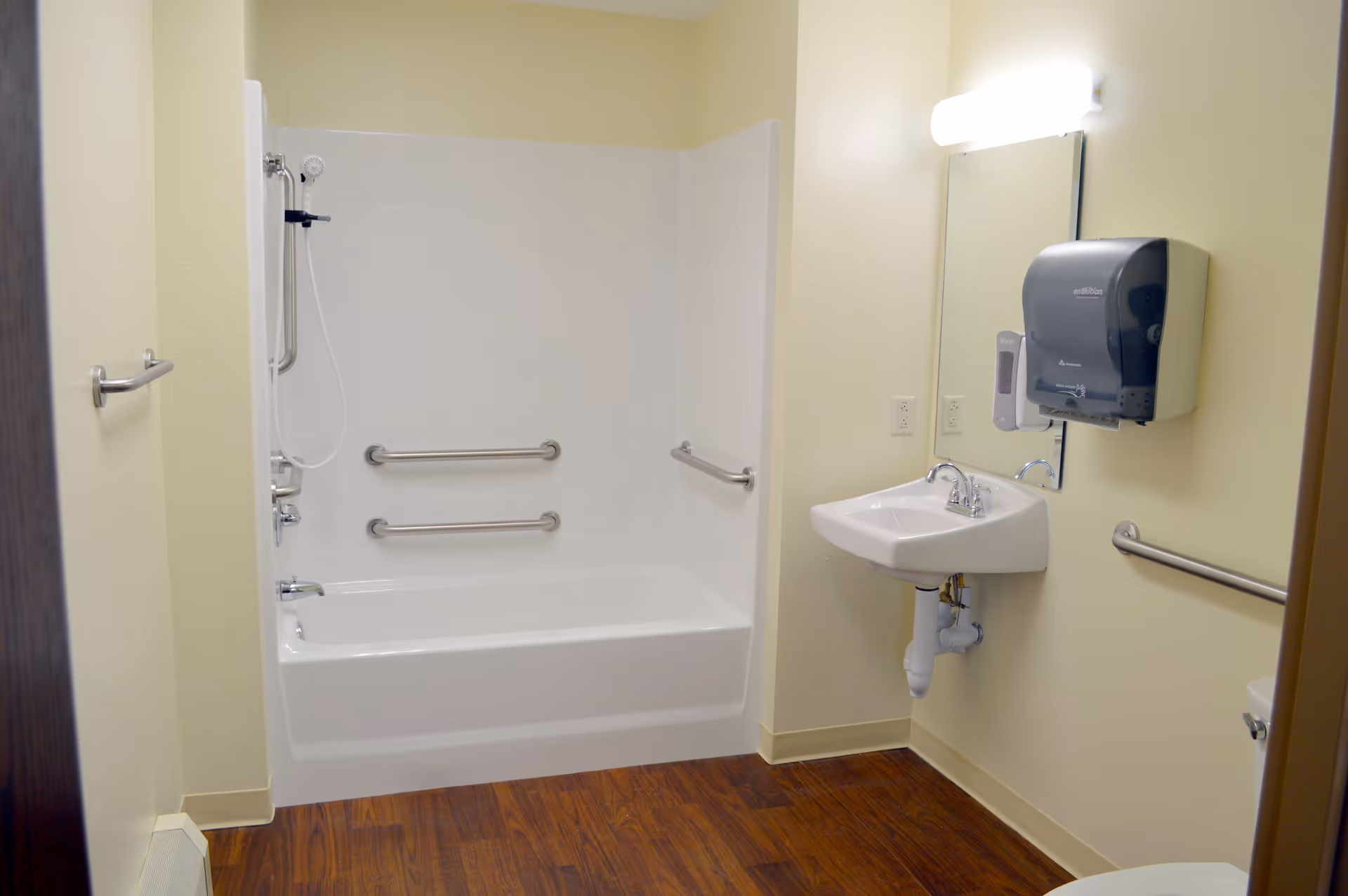 Accessible bathroom with a bathtub/shower, multiple grab bars, wall-mounted sink and mirror, and a soap dispenser.